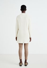 Long gilet tricoté de couleur crème avec une texture côtelée, porté sur des leggings beiges et des chaussures à enfiler à motif animal. Coupe simple et décontractée.