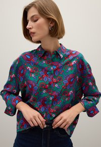 Frau mit kurzen Haaren, die silberne Creolen und ein lebhaft grünes Hemd mit rotem, blauem und pinkem blumigen Paisley-Muster trägt.