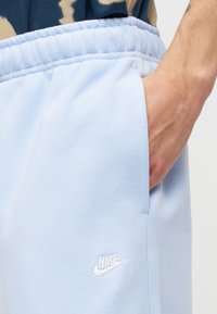 Main reposant dans la poche d'un pantalon de survêtement Nike bleu clair avec logo blanc brodé, taille élastique, et partie d'une chemise à motifs visible.