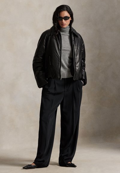 Jachetă bomber din piele neagră purtată peste un pulover gri cu guler înalt, asortată cu pantaloni largi negri și pantofi negri fără toc. Modelul poartă ochelari de soare întunecați.