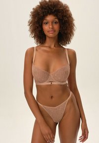 Beżowy komplet braletki i stringów z koronkowymi wzorami, regulowane ramiączka oraz akcent z przodu z zapięciem. Miękka tekstura i delikatny design.