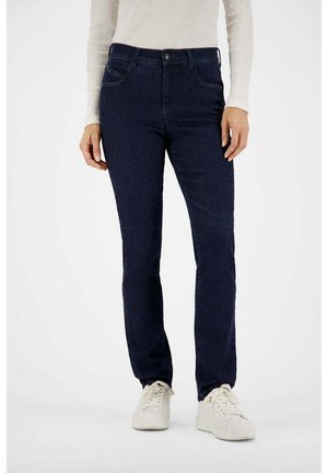 Frau trägt dunkelblaue Jeans mit geradem Bein, weißes langärmeliges Oberteil und weiße Sneakers, steht vor einem schlichten Hintergrund.