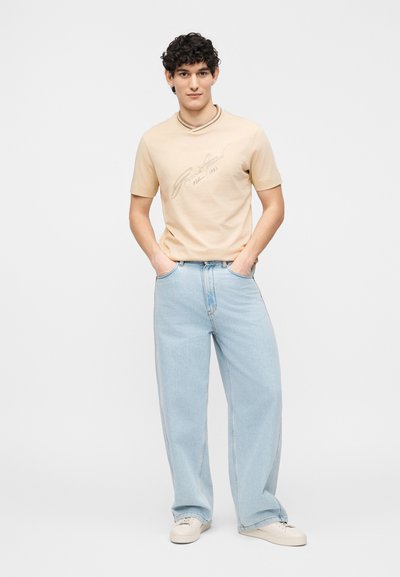 Άνδρας με κατσαρά μαλλιά στέκεται φορώντας ένα μπεζ κοντομάνικο πουκάμισο με κείμενο, ανοιχτό μπλε τζιν με φαρδιά μπατζάκια και λευκά αθλητικά, με τα χέρια στις τσέπες.