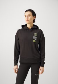 Schwarzer Hoodie mit einer Vordertasche und Bündchen, ausgestattet mit grünen und weißen Logos und Text. Glatte Textur, lockere Passform, kombiniert mit schwarzen Leggings.