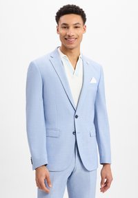 Hellblauer Blazer aus strukturiertem Stoff, mit zwei Knöpfen, Reverskragen und einem weißen Taschenquadratl. Über einem hellen Hemd getragen.