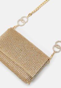 Torba w złotym odcieniu, pokryta małymi kryształkami, o prostokątnym kształcie, z łańcuszkowym paskiem i okrągłymi ozdobami z przezroczystymi kryształami.