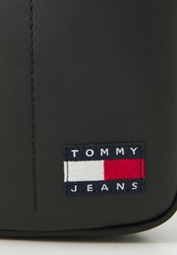 Svart faux skinnveske med en søm og et rektangulært brodert merkefelt med røde, hvite og blå detaljer og teksten "TOMMY JEANS".