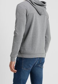 Persona con indosso una felpa grigia con cappuccio e jeans blu, mostrata di spalle contro uno sfondo chiaro e uniforme.