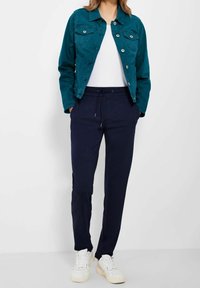 Teal denimová bunda s dvěma náprsními kapsami, bílý tričko, námořnické teplákové kalhoty s elastickým pasem a bílé tenisky. Hladká textura, volný střih.