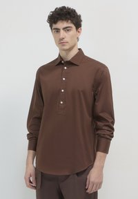 Junger Mann mit kurzen lockigen Haaren, der ein langärmliges braunes Hemd mit Knopfleiste trägt, steht vor einem schlichten hellen Hintergrund.