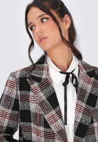 Donna con capelli scuri, indossante un blazer a quadri nero, rosso e bianco, una camicia bianca con colletto, una collana di perle e una cravatta nera, che guarda di lato.