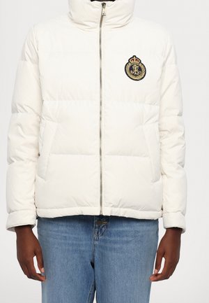 Witte pufferjack met ritssluiting aan de voorkant en geborduurd wapen op de borst, gedragen over een blauwe spijkerbroek, gemodelleerd door een persoon met ontspannen handen langs het lichaam.