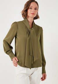 Olive green blouse in smooth fabric with a bow tie collar, long puffed sleeves, and a rounded hem, paired with white trousers.