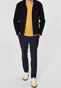 Marineblauwe wollen jas met een zwarte ritssluiting, gele gebreide top eronder, marineblauwe broek en crèmekleurige sneakers met bruine zolen. Casual outfit indeling.