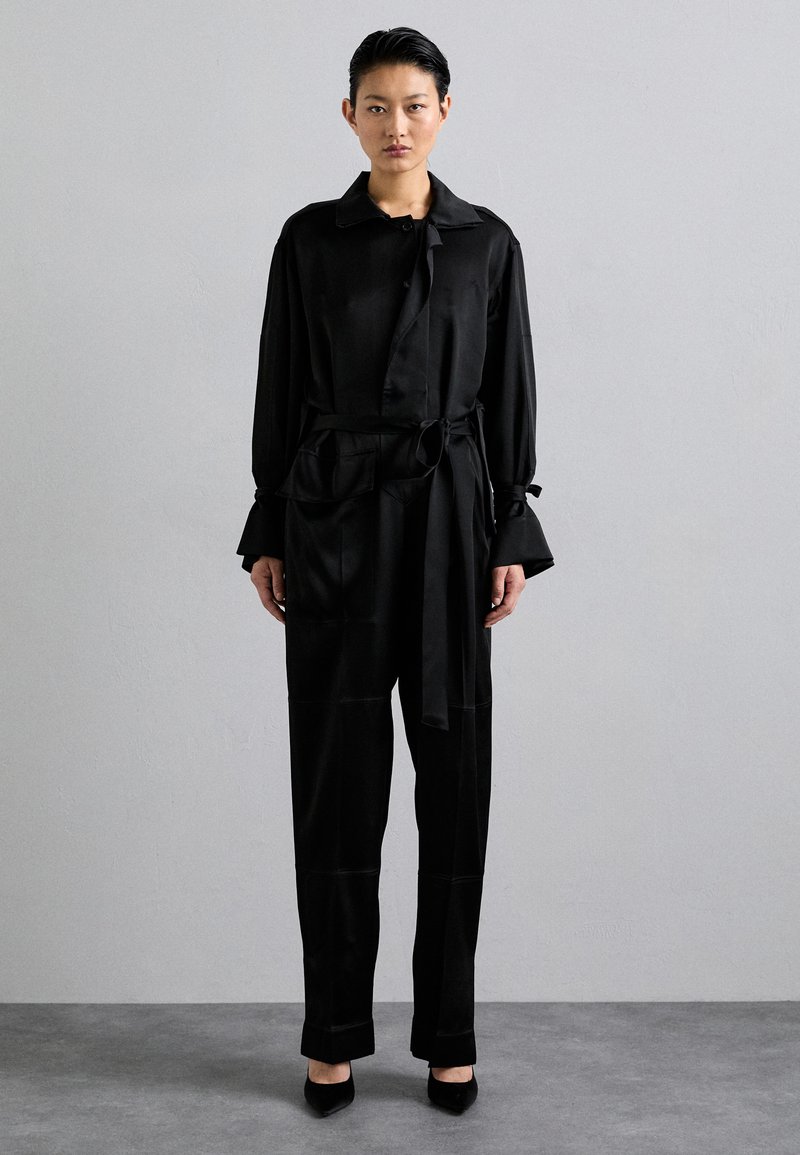 Conjunto de satén negro que incluye una blusa de manga larga con cuello, cinturilla atada y pantalones de pierna ancha. Textura suave con bolsillos en la parte delantera.