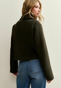 Vrouw met blond haar, gekleed in een cropped donkergroen jasje en hooglopende blauwe spijkerbroeken, staand tegen een effen lichte muur, in zijaanzicht.