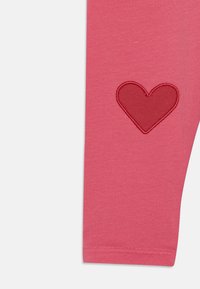 Rozā legingi ar mīkstu tekstūru, kuros kreisajā pusē ir sarkans izšūts sirds dizains. Vienkārša forma ar šūtu malu.