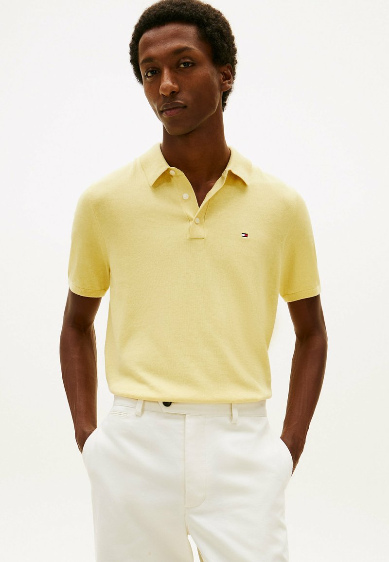 Homme portant un polo à manches courtes de couleur jaune pâle et un pantalon blanc, debout les mains dans les poches devant un arrière-plan uni.