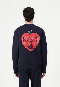 Marineblå sweatshirt med et stort rødt æblegrafik på ryggen, med teksten "LES DEUX LEGACY COURT" og et grønt blad. Blødt, afslappet stof.