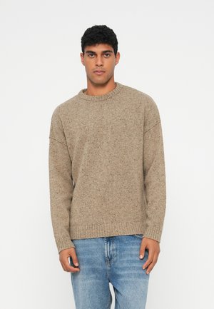 Beige, stickad tröja med rund halsringning, ribbade ärmslut och nederkant. Texturen är mjuk med ett subtilt prickigt mönster, i kombination med ljusblå jeans.