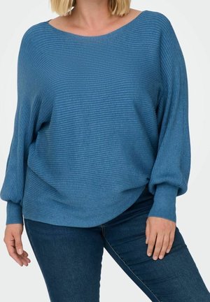 Femme portant un pull bleu côtelé ample à manches longues et un jean bleu foncé, debout avec une main posée sur sa cuisse.