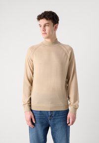 Beige langermet genser med høy hals, laget av mykt stoff. Har raglanermer og ribbestrikkede mansjetter og fald. Kombinert med blå jeans.
