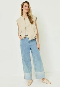 Donna che indossa un gilet smanicato beige con cerniera, top bianco, jeans a gamba larga azzurri con orli risvoltati, sneakers beige e un orologio da polso, in posizione rilassata.