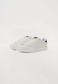 Hvite lær-sneakers med rund tå, teksturert overflate, flate snørebånd og en kontrastfarge i marineblå hælflik. Minimalistisk design med logo.