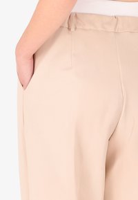 Pantaloni beige con vestibilità rilassata, texture liscia, tasche laterali e un design semplice della vita. Nessun motivo visibile o accessori metallici.