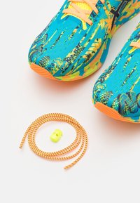 Chaussures de course bleu vif et orange avec tissu à motifs et lacets orange à côté d'un lacet élastique orange roulé avec fermeture jaune.