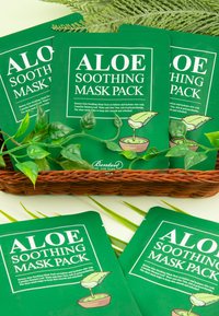 Packaging verde per la "Maschera Idratante Aloe", con grafica della pianta di aloe e testo bianco, sistemato in un cestino di vimini con foglie verdi.