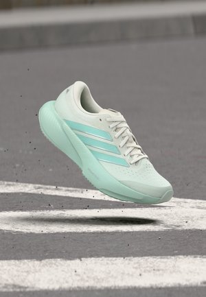 Chaussure de course bleu sarcelle clair et blanche avec trois bandes flottant au-dessus d'une route avec des marquages blancs.