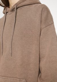 Brązowa bluza z kapturem wykonana z miękkiego materiału, z przednią kieszenią, sznurkami oraz haftowanym napisem „BDG JEANS” w subtelnym różowym odcieniu.