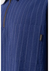 Modrý texturovaný látkový top s zipovým výstřihem, vertikálními bílými proužky a malou nášivkou na boku. Obsahuje dvě náprsní kapsy.