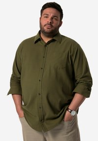 Camicetta verde oliva con taschino, bottoni neri e maniche arrotolate, abbinata a pantaloni beige chiaro e un orologio marrone.