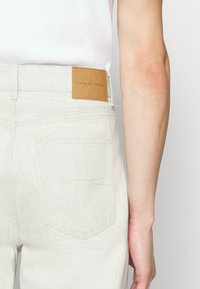 Pantalones claros de mezclilla, con un parche de cuero etiquetado "TIGER OF SWEDEN" en la cinturilla y dos bolsillos traseros.