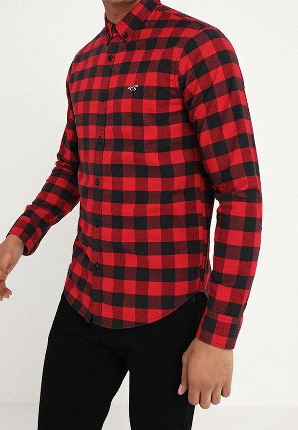 Camisa de franela a cuadros rojos y negros con cuello abotonado, mangas largas y acento de logo en el lado izquierdo del pecho. Dobladillo ligeramente curvado.