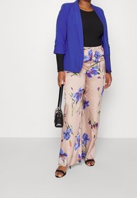 Mujer con blazer azul, top negro, pantalones anchos beige con flores, tacones negros, sosteniendo un pequeño bolso negro.