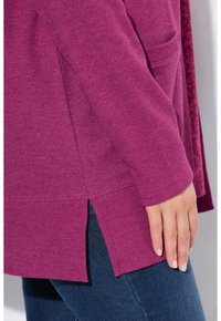 Fuchsia Strickjacke mit gerippter Textur, seitlichen Schlitzen und einer Vordertasche. Über dunkler Denim getragen. Weicher Stoff für Komfort.