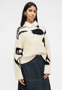 Γυναίκα που φοράει κρεμ πουλόβερ με μαύρα αφηρημένα σχέδια πάνω από λευκό πουκάμισο με γιακά και σκούρα φούστα από τζιν, στέκεται μπροστά σε απλό φόντο.