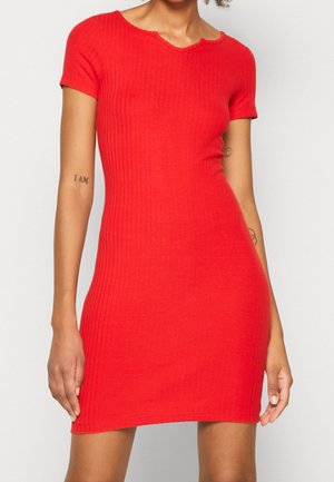 Frau trägt ein eng anliegendes, kurzärmeliges, geripptes rotes Kleid und steht vor einem schlichten, hellen Hintergrund.