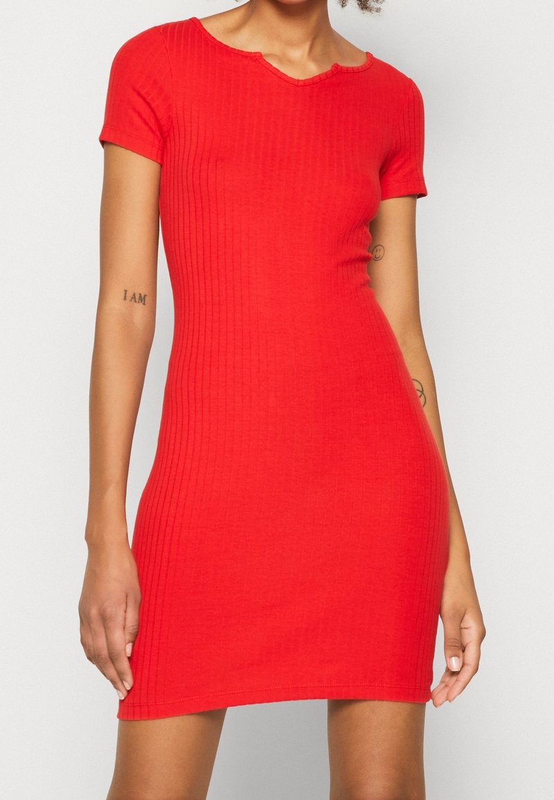 Femme portant une robe rouge moulante à manches courtes côtelée, debout devant un fond clair uni.