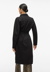 Cappotto lungo nero con vita regolabile, spalle strutturate e colletto. Il materiale sembra liscio; presenta una cucitura sul retro e una silhouette dritta.