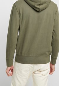 Sweat à capuche en coton vert olive avec capuche à cordon et poignets côtelés. Présente un design simple sans motifs ni logos visibles.