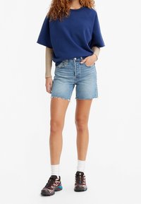 Blå kortärmad sweatshirt i kombination med ljusa denimshorts som har fransade kanter och en avslappnad passform, kompletterad med sportiga svarta och blå sneakers.