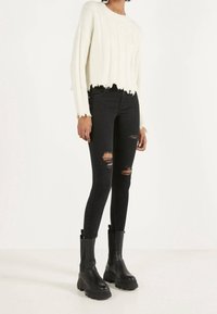 Suéter blanco, recortado, con textura acanalada y dobladillo deshilachado, combinado con jeans negros ajustados con detalles rasgados, y botas negras gruesas.