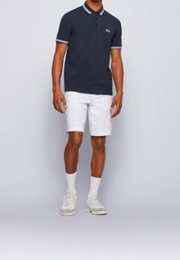 Polo shirt navy avec col rayé, shorts blancs, chaussettes blanches mi-mollet et baskets de couleur claire. Présente un petit logo sur la poitrine.