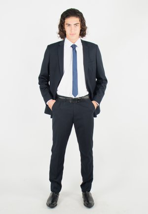 Costume bleu marine avec une chemise blanche et une cravate bleue. Coupe ajustée, tissu lisse, avec une poche sur la veste. Chaussures en cuir noir.