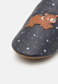 Pantoufle en cuir marine avec des motifs géométriques en bleu clair et marron. Présente un appliqué de ours marron brodé sur le dessus. Bout doux et arrondi.