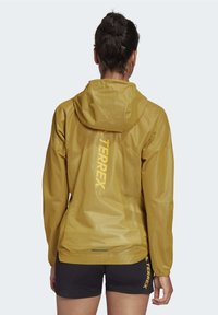 Gele waterdichte jas met capuchon en elastische manchetten, met het "TERREX"-logo verticaal op de achterkant gedrukt. Lichtgewicht, gladde textuur.